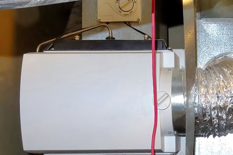 A close-up view of a whole-house bypass humidifier unit mounted on metal HVAC ductwork, connected to flexible foil ducting on the right and a control dial above.