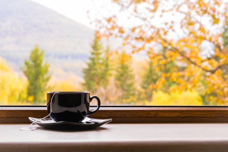 A black mug of tea resting on a windowsill, looking out onto a scenic view of colorful autumn trees and mountains.